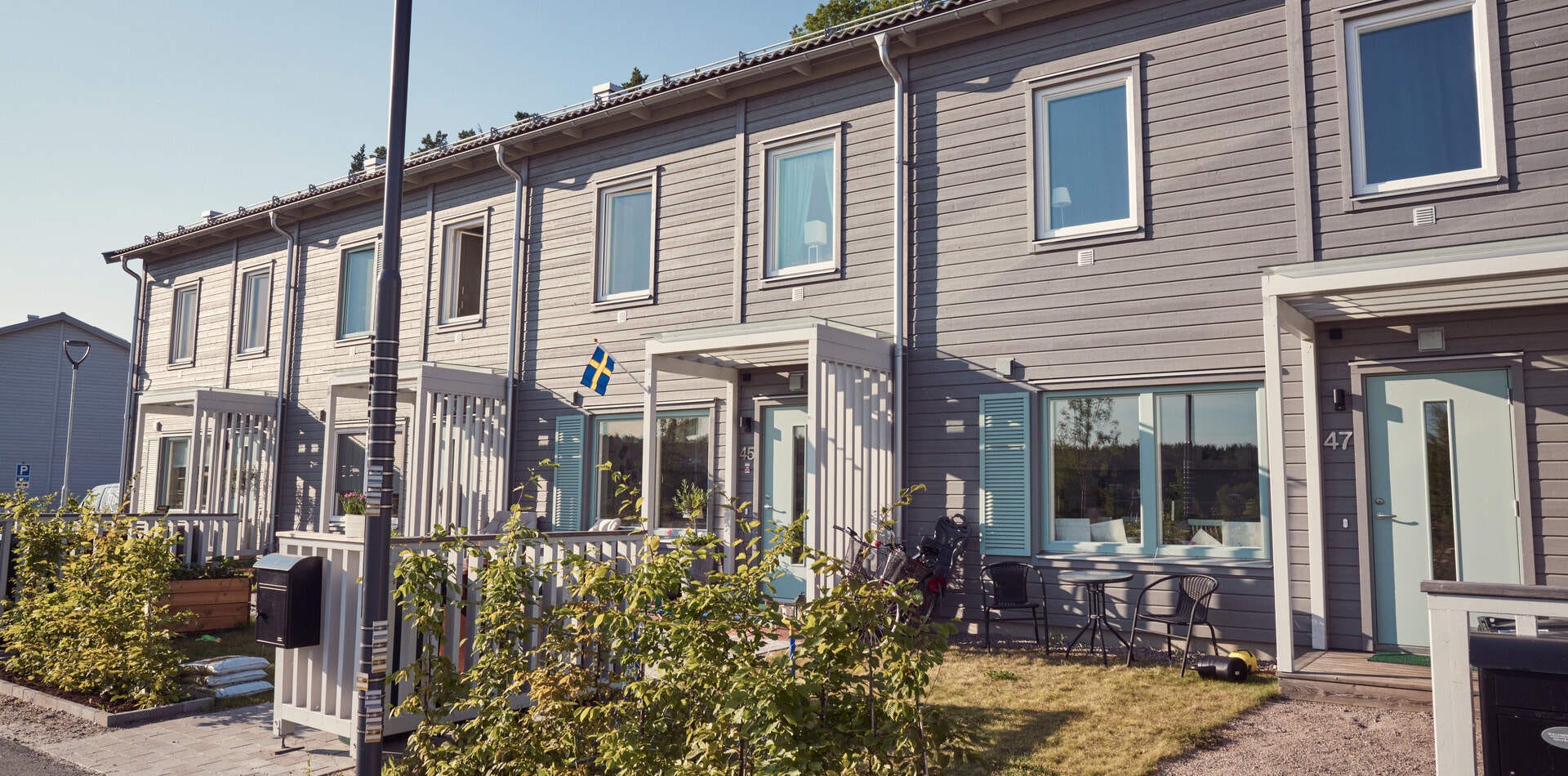 Foto från tidigare liknande grannskap Foto - Radhus i två våningar med grå fasad. Varje enhet har en egen ingång med liten veranda. En svensk flagga hänger vid ingången.