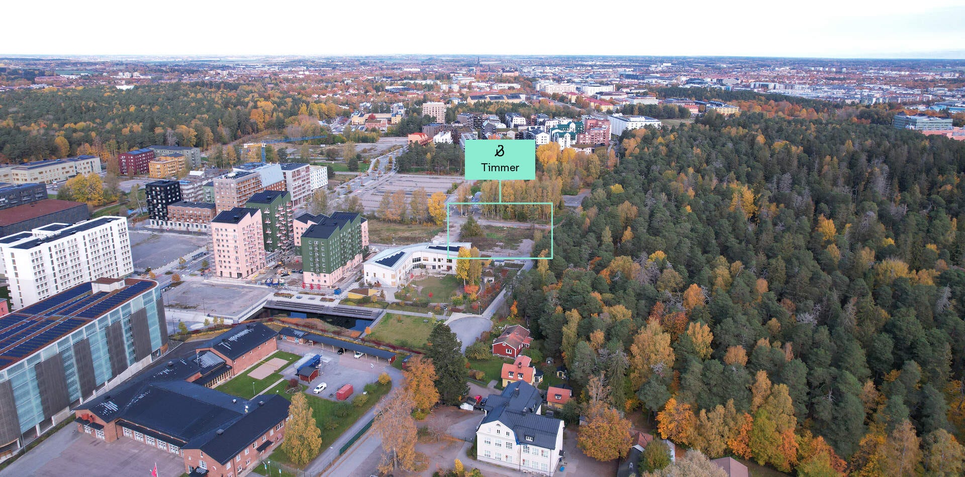 Foto - Nyproduktion i Rosendal, Uppsala - Flygfoto över stadsmiljö med skog. Texten "Timmer" markerar en plats. Färgglada byggnader omges av höstfärger.