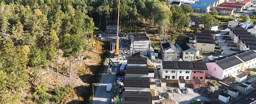 Flygbild över ett nybyggt eller pågående bostadsområde med radhuslängor, byggarbetsplats och skogsområde intill.
