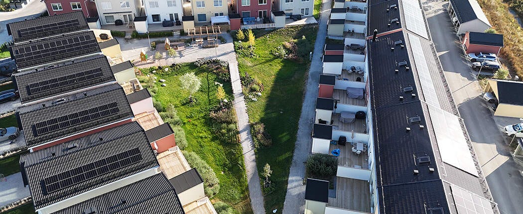 Flygfoto över ett radhus-/kvartershusområde med gemensam gård, gångstråk och långa takytor med solceller.