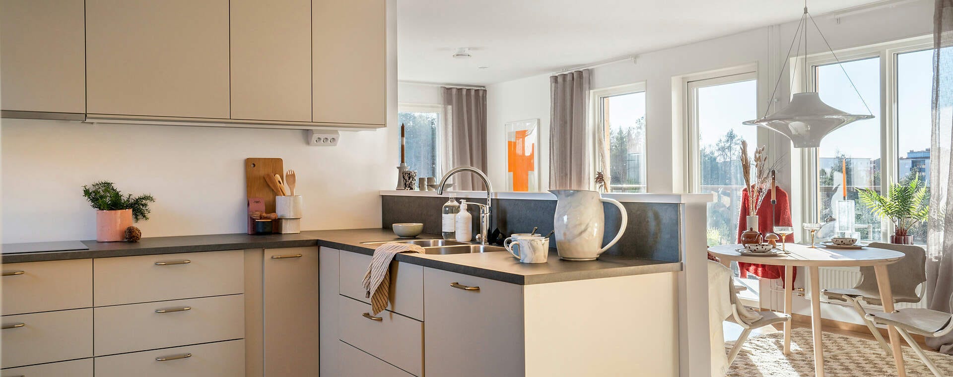 Foto - Ljust kök med grå bänkskiva, diskho och matplats vid stora fönster med dagsljus och modern inredning.